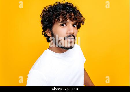 Close up of confused puzzled curly haired indian or arabian guy in white t-shirt, looking questioningly at the camera, while standing over isolated orange background Stock Photo