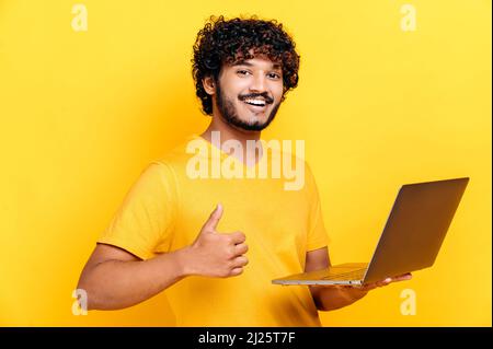 Happy arabian or indian guy with curly hair, wearing a t-shirt, holding an open laptop in his hand, looking at the camera with smile, showing thumbs up gesture, standing on isolated orange background Stock Photo