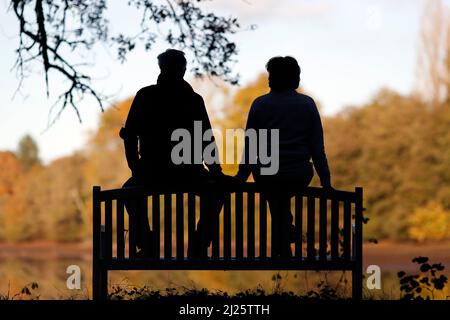 Silhouettes of woman and man sitting on bench against lake and forest. Stock Photo