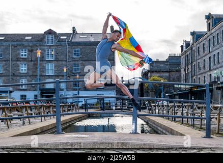 Leith, Edinburgh, UK, Scotland, 30 March 2022. Edinburgh International Festival launch: Scottish Ballet principal dancer Jerome Anthony Barnes performs a leap into the air with the anniversary flag to celebrate the 75th anniversary of the festival in Commercial Street Stock Photo