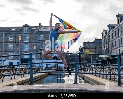 Leith, Edinburgh, UK, Scotland, 30 March 2022. Edinburgh International Festival launch: Scottish Ballet principal dancer Jerome Anthony Barnes performs a leap into the air with the anniversary flag to celebrate the 75th anniversary of the festival in Commercial Street Stock Photo