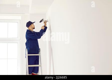 Handyman in overalls installs surveillance camera on wall mounting inside new house. Stock Photo