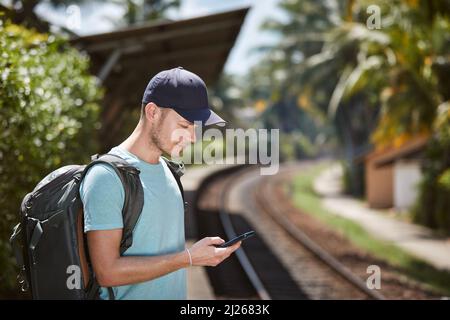 Tourist with backpack in tropical travel destination. Side view of man using mobile phone while waiting for train at railroad station. Stock Photo