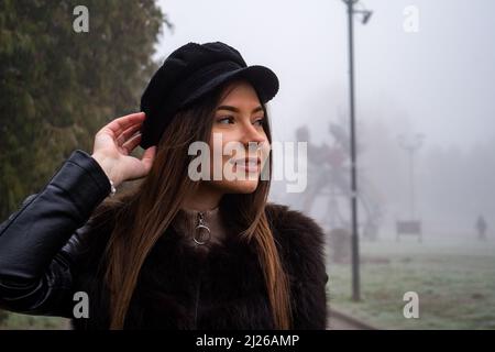 Smiling girl in trench coat walks in park with bouquet of autumn leaves on foggy morning. Stock Photo