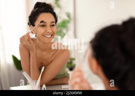Charming millennial brunette female in towel applying eyebrow mascara near mirror after bath at home, copy space Stock Photo