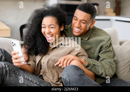 Overjoyed couple resting on couch and holding smartphone, while celebrating on-line lottery win,bid betting victory moment, unbelievable opportunity or invitation, internet sale, getting prize concept Stock Photo