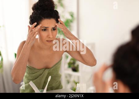 Concerned millennial female looking at her reflection in mirror, checking first wrinkles on forehead at bathroom, free space Stock Photo