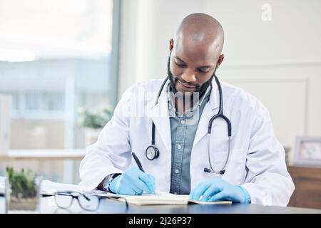 The research never ends, even when youre qualified. Shot of a handsome young doctor sitting alone in his clinic and making notes. Stock Photo