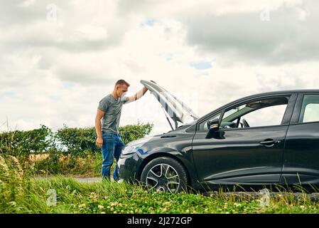 Nobody wants their journey cut short by car problems. Shot of a young man checking under the hood of his car after breaking down on the road. Stock Photo