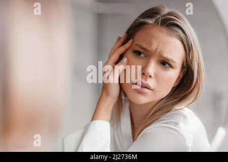 Acne prone skin concept. Sad lady looking at mirror and checking her face skin, standing in bathroom interior Stock Photo