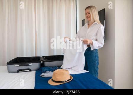 Young happy blonde woman is holding a white T-shirt in her hands, putting things in a suitcase. Concept of travel, recreation, tourism Stock Photo