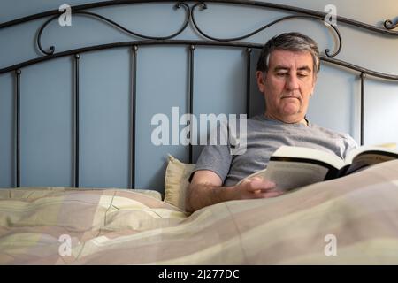 Man with insomnia sitting on the bed and reading a book. Stock Photo
