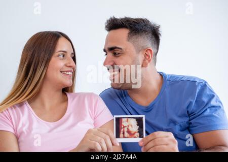 portrait of happy pregnant heterosexual caucasian young couple laughing inside bedroom looking at each other and showing ultrasound printout of their Stock Photo