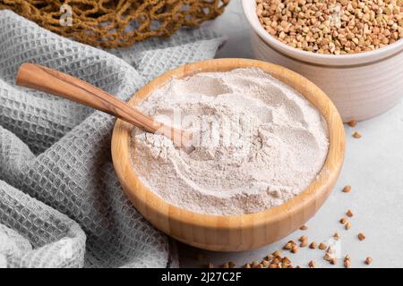 Buckwheat flour in a wooden bowl with wooden spoon and raw green buckwheat grain in a bowl on light grey stone background, close up. Alternative flour Stock Photo