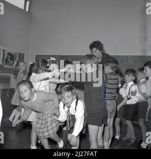 1960s, historical, primary school children playing a singing game in the classroom, possibly the one called 'Oranges and Lemons', where children move or dance through an arch made by children holding hands, facing each other, arms raised, Fife, Scotland, UK. The game is also known as 'London Bridge is Falling Down', where again two or more children join hands to form an arch, the bridge, where the other choldren march under. In Scotland it is also known as 'broken bridges'. Stock Photo