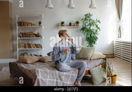 a young man is talking on a mobile phone, sitting on the edge of the bed with a cup of coffee Stock Photo