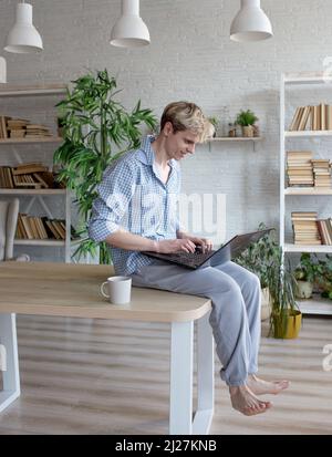 A young programmer guy is sitting on top of a table with his legs hanging down while working on a laptop. Home environment, freelancer Stock Photo