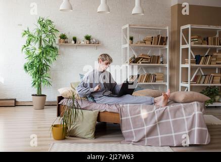 Smiling handsome young man 20 years old, working on a laptop computer, lying in bed, resting, spending time in the bedroom at home Stock Photo