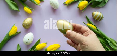 Banner with Easter greetings: close up hand holds golden egg, yellow tulips, painted white eggs lilac background. Copy space.