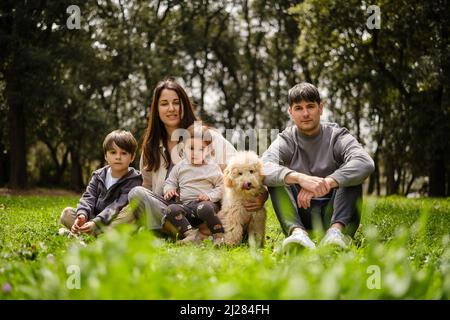 Portrait of a family and dog sitting together on the grass while enjoying the nature outdoors. Stock Photo