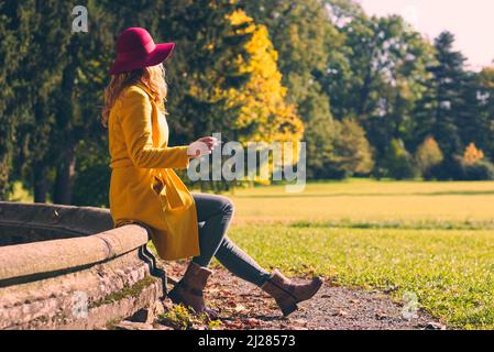 Fashionable woman sitting on old fountain and enjoying autumn season and smoking cigarette Stock Photo