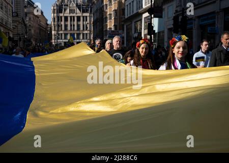 Young women with traditional flowers in their hair hold a giant Ukrainian flag as thousands of people gather in the capital for a peace march ‘London stands with Ukraine’ in solidarity with the people of Ukraine on 26th March 2022 in London, United Kingdom. The conflict in Ukraine is now in it’s second month and their president has called for people across the World to unite in protest at the Russian invasion and ongoing war in the region. The Ukrainian flag of yellow and blue was everywhere to be seen in this colourful show of unity. Stock Photo