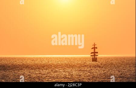 A red burning sunset with the silhouette of sailing ship. Sailors set sails on masts of ship. White sailing vessel floating in the sea. Copy space. Stock Photo