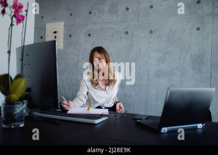 The Caucasian businesswoman is sitting in her grey interior office, in a cozy environment. A beautiful middle-aged woman is using a laptop and desktop Stock Photo