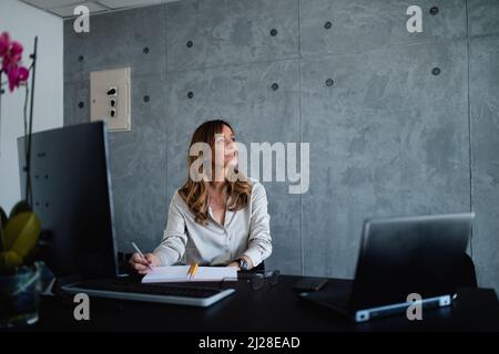 The Caucasian middle-aged female manager is thinking about employees and how to compose a schedule in her favorite beautiful, grey office. Stock Photo