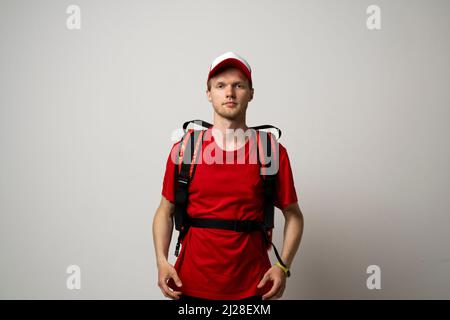 Young courier in a red uniform t-shirt and with red food thermo bag on a shoulder standing isolated on white background studio. Food delivery service. Stock Photo