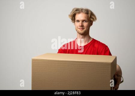 Delivery service. Young smiling courier holding cardboard box. Happy young delivery man in red t-shirt standing with parcel isolated on white Stock Photo