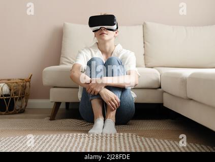 Testing new technologies. Attractive young woman in VR headset gesturing and smiling while sitting at home. Woman in VR headset looking up at the obje Stock Photo