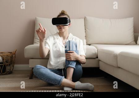 Testing new technologies. Attractive young woman in VR headset gesturing and smiling while sitting at home. Woman in VR headset looking up at the obje Stock Photo