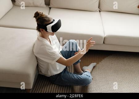 Testing new technologies. Attractive young woman in VR headset gesturing and smiling while sitting at home. Woman in VR headset looking up at the obje Stock Photo