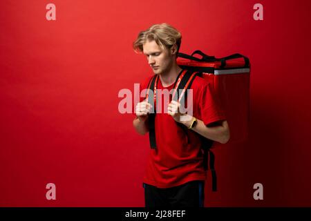 Young delivery employee man in red tshirt uniform with a thermal food bag backpack works as a courier in a food delivery service on a white background Stock Photo