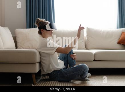 Testing new technologies. Attractive young woman in VR headset gesturing and smiling while sitting at home. Woman in VR headset looking up at the obje Stock Photo