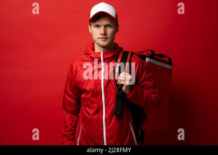 Young courier in a red uniform t-shirt and with red food thermo bag on a shoulder standing isolated on red background studio. Food delivery service. Stock Photo