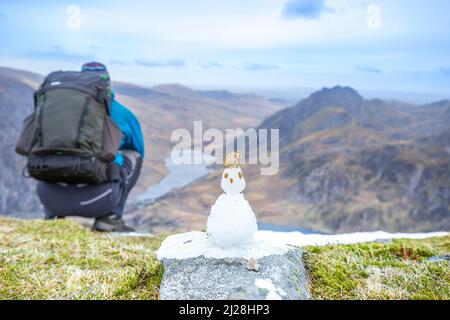 Rear view of male hiker & funny mini snowman on top of Y Garn mountain overlooking Tryfan & Lake Ogwen, Snowdonia National Park, North Wales, UK. Stock Photo