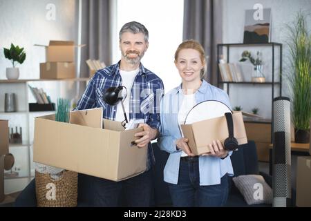 Caucasian middle aged couple in casual wear smiling and hugging at new flat, holding boxes with different decor and things. Young family moving to modern apartment. Cardboxes around. Stock Photo