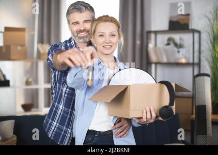 Middle aged Caucasian family, bearded man and blond lady, standing at new house with box and and showing keys on camera. Excited man and woman hugging and smiling indoors. Stock Photo