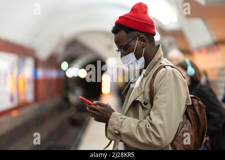 African man in face mask in subway station wait for train, using smartphone, scrolling social media Stock Photo
