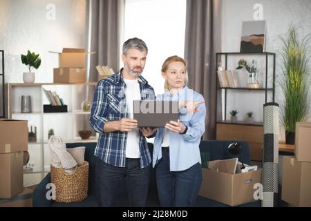 Caucasian mature couple standing among cardboxes and different things and using laptop, looking for the design and making online shopping. Family taking break during removing to new flat. Stock Photo