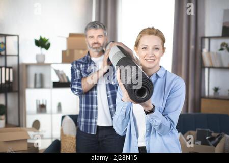 Portrait of a smiling middle aged couple carrying rolled rug after moving in a house. Focus on smiling pretty blond woman, holding roll carpet on her shoulder and her pleasant bearded man behind Stock Photo