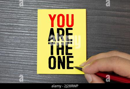 A man is holding a piece of paper which is saying 'You are the one '. The paper is white with black handwritten letters and the background is red. Stock Photo