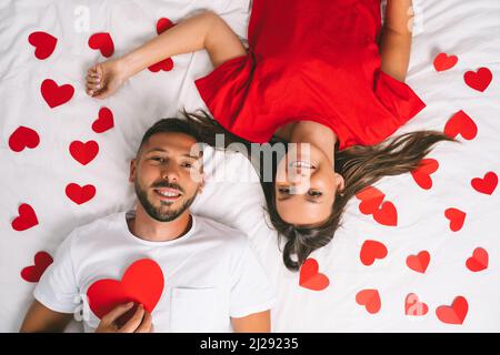 Top view of happy young couple in love with red paper hearts lying on the white bed. Love and relationship Stock Photo