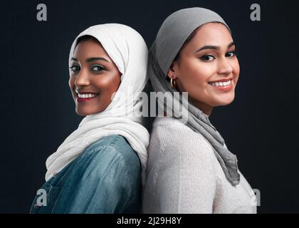 We will always have each others backs. Cropped shot of two attractive young women wearing hijabs and standing back to back against a black studio Stock Photo