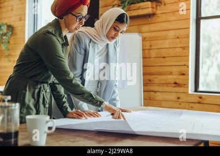 Innovative female architects working on blueprint drawings in a modern office. Two Muslim businesswomen planning a new creative project. Two designers