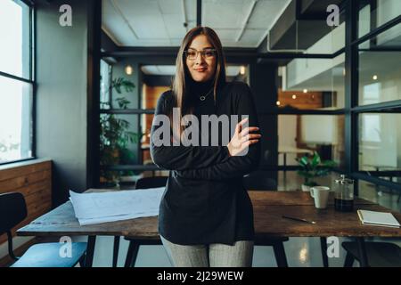 Young female entrepreneur looking at the camera while standing with her arms crossed. Cheerful young businesswoman standing in the boardroom of a mode Stock Photo