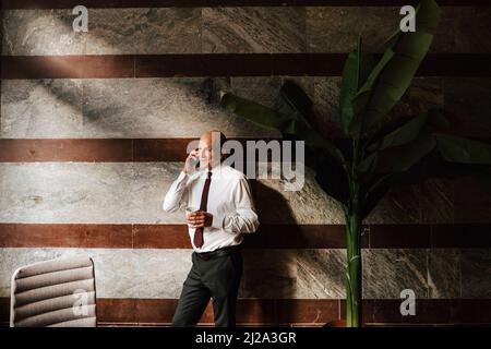 Smiling senior businessman holding coffee cup talking through mobile phone leaning on wall at office Stock Photo