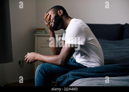 Emotionally stressed man covering eyes with hand sitting on bed at home Stock Photo